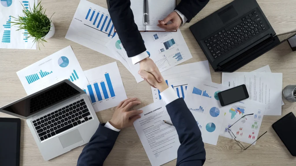two people shaking hands over a table with scattered papers showing graphs and open laptops